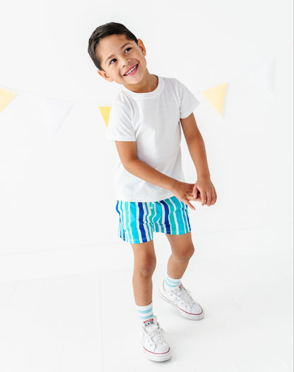 A young boy smiling and posing in a white t-shirt, blue and green striped shorts, white socks, and white sneakers against a bright white background with a subtle yellow bunting banner.