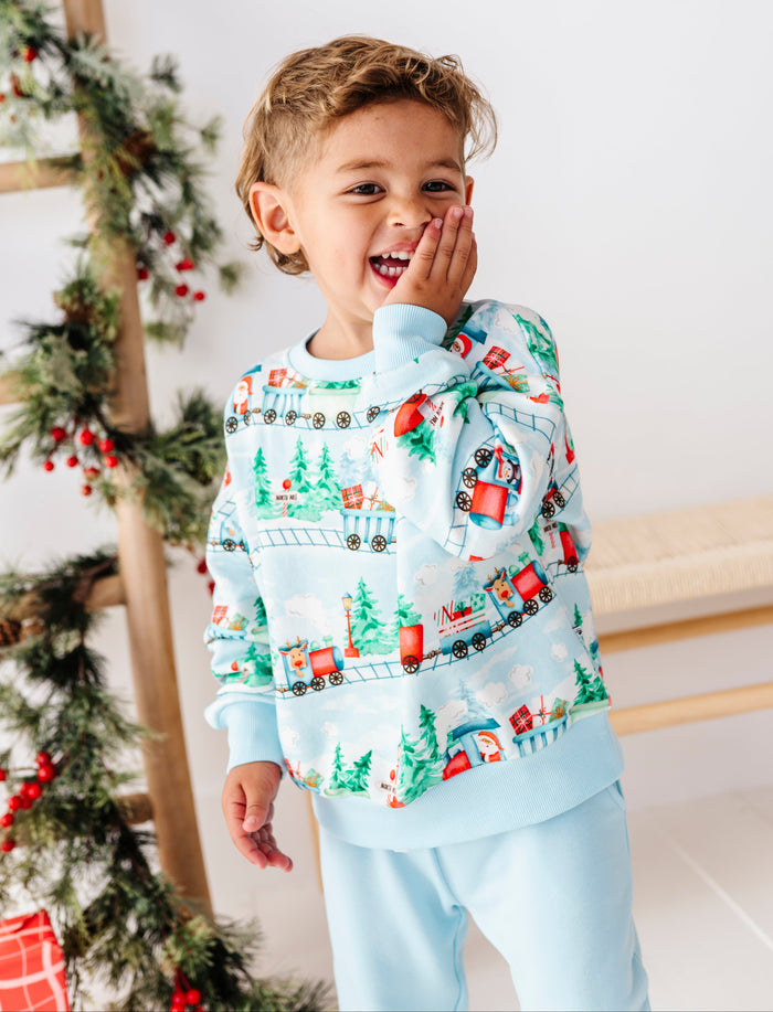 A young boy wearing a light blue Christmas-themed long-sleeve crewneck top and matching sweatpants, standing in a festive indoor setting with a decorated Christmas tree on the left.