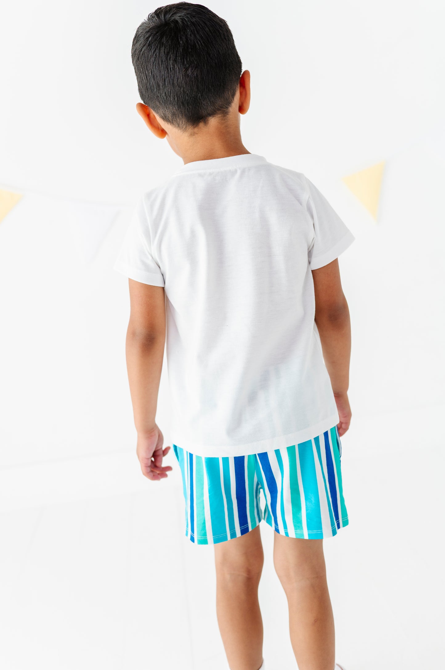 A child viewed from behind wearing a plain white short-sleeve T-shirt and bright blue and white vertically striped shorts against a clean white background.