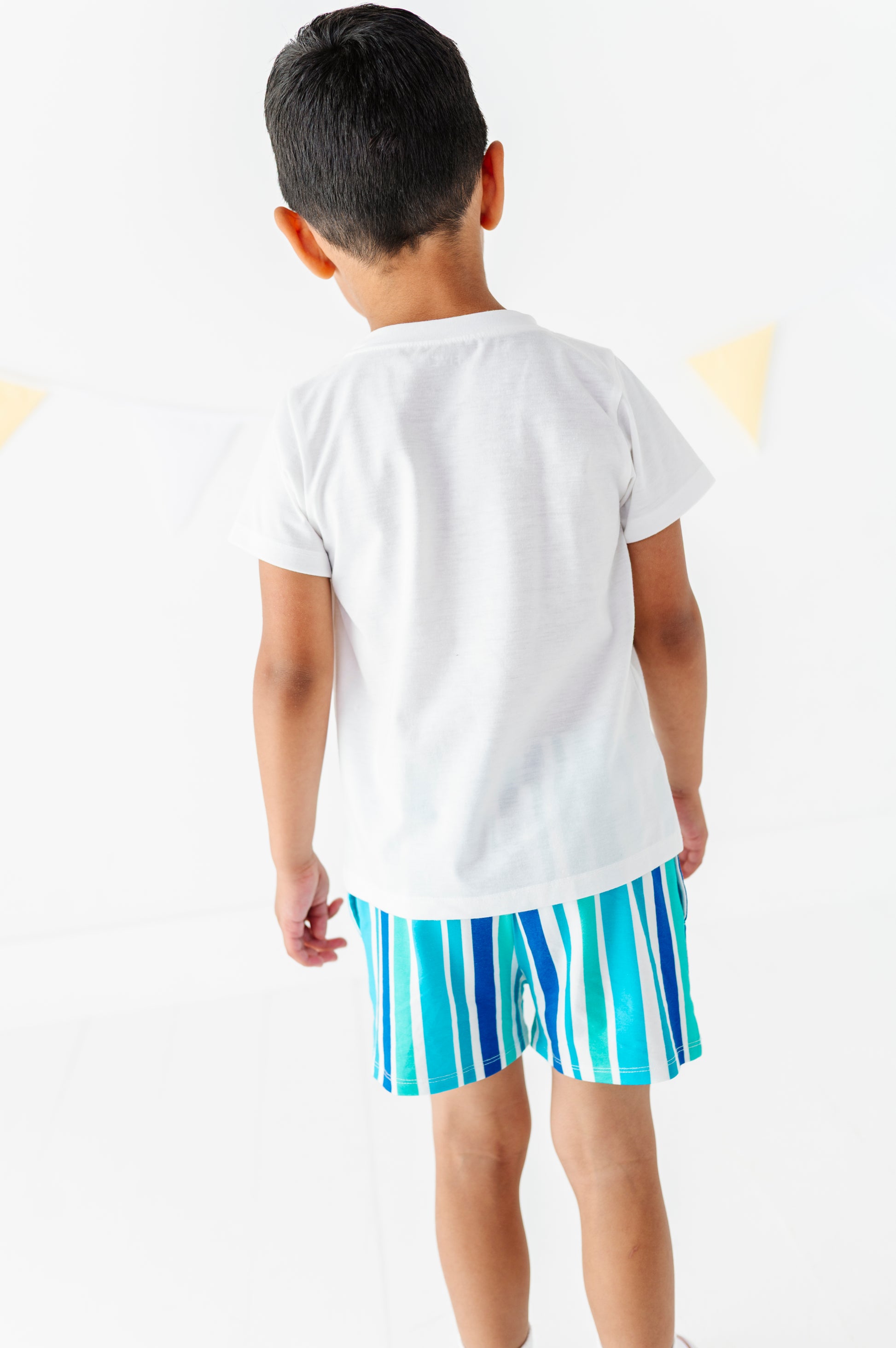 A child viewed from behind wearing a plain white short-sleeve T-shirt and bright blue and white vertically striped shorts against a clean white background.