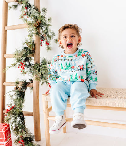 A young child wearing a festive Christmas sweater and light blue sweatpants sits excitedly on a wooden bench, with a decorated wooden ladder and holiday garland to the left.