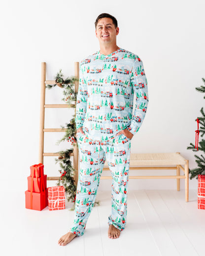 A man wearing a matching long-sleeve bamboo pajama set with a Christmas tree and gift pattern, standing barefoot in a bright room with a wooden bench, ladder, and wrapped presents.