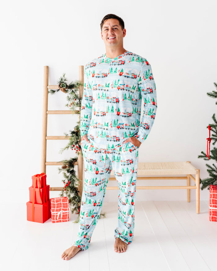 A man wearing a matching long-sleeve bamboo pajama set with a Christmas tree and gift pattern, standing barefoot in a bright room with a wooden bench, ladder, and wrapped presents.