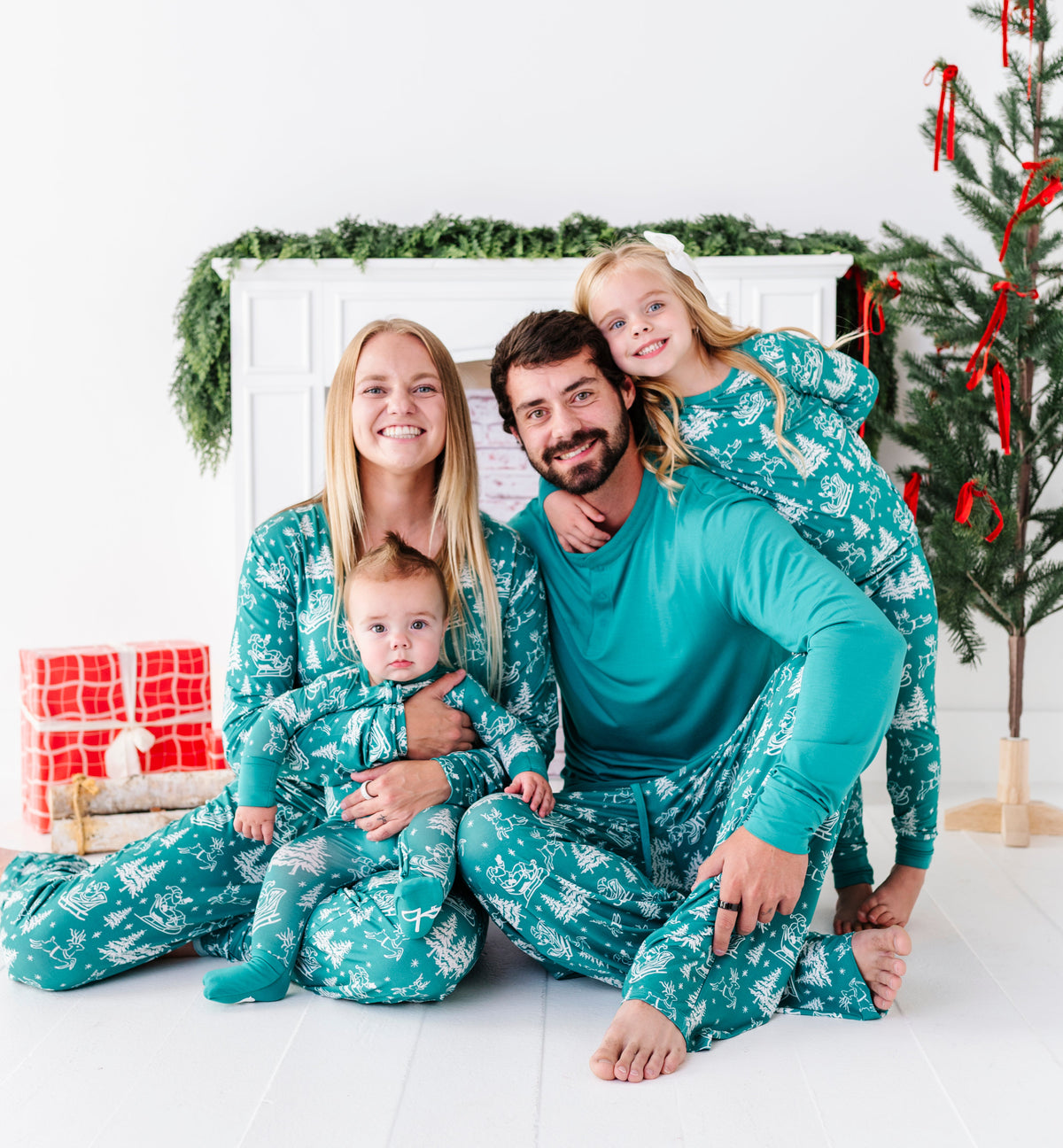 A family photo sitting in front of a fireplace and a Christmas tree. The mom, dad, daughter, and baby are all wearing matching bamboo pajamas featuring a vintage Christmas print 