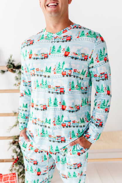 A man wearing a light blue long-sleeve bamboo pajama top and matching pants with a repeating Christmas train and pine tree pattern, standing indoors near a decorated holiday tree.