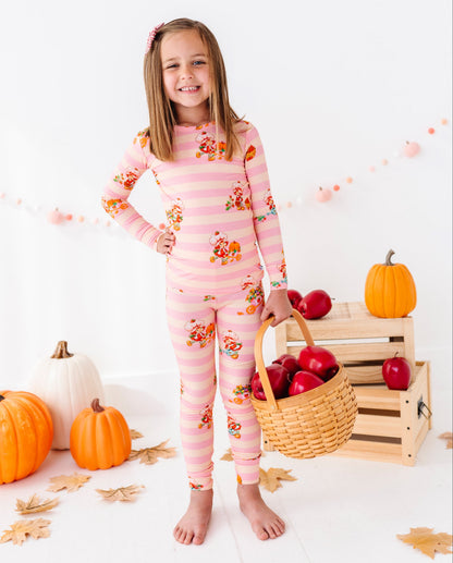 A young girl stands barefoot in a studio setting wearing a pink long-sleeve pajama set with horizontal stripes and a small floral print. She holds a wicker basket filled with red apples next to stacked wooden crates holding more apples and pumpkins.