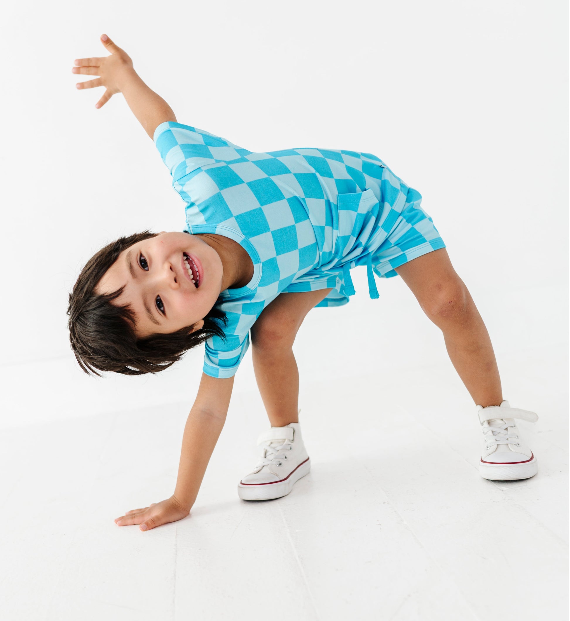 A young child wearing a blue checkered romper and white sneakers poses on a white studio floor against a white background.