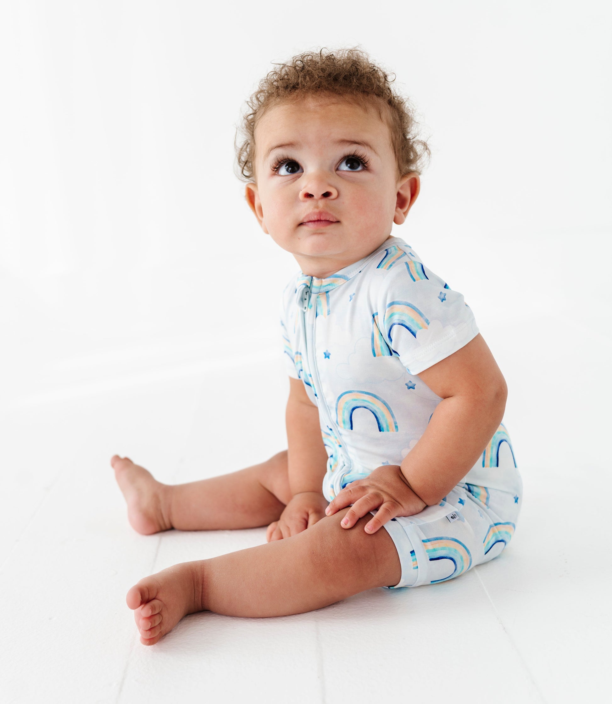 Baby wearing a light blue shortie zip romper with rainbow patterns on a white background