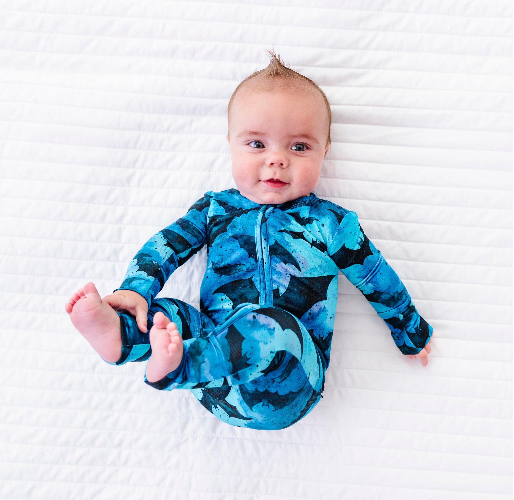 A baby lying on a white textured blanket, wearing a long-sleeve zip-up onesie with a blue and black abstract pattern.
