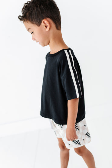 A young boy wearing a black short-sleeve t-shirt with white double stripes down the sleeves, paired with white shorts featuring a black graphic pattern, set against a clean white background.