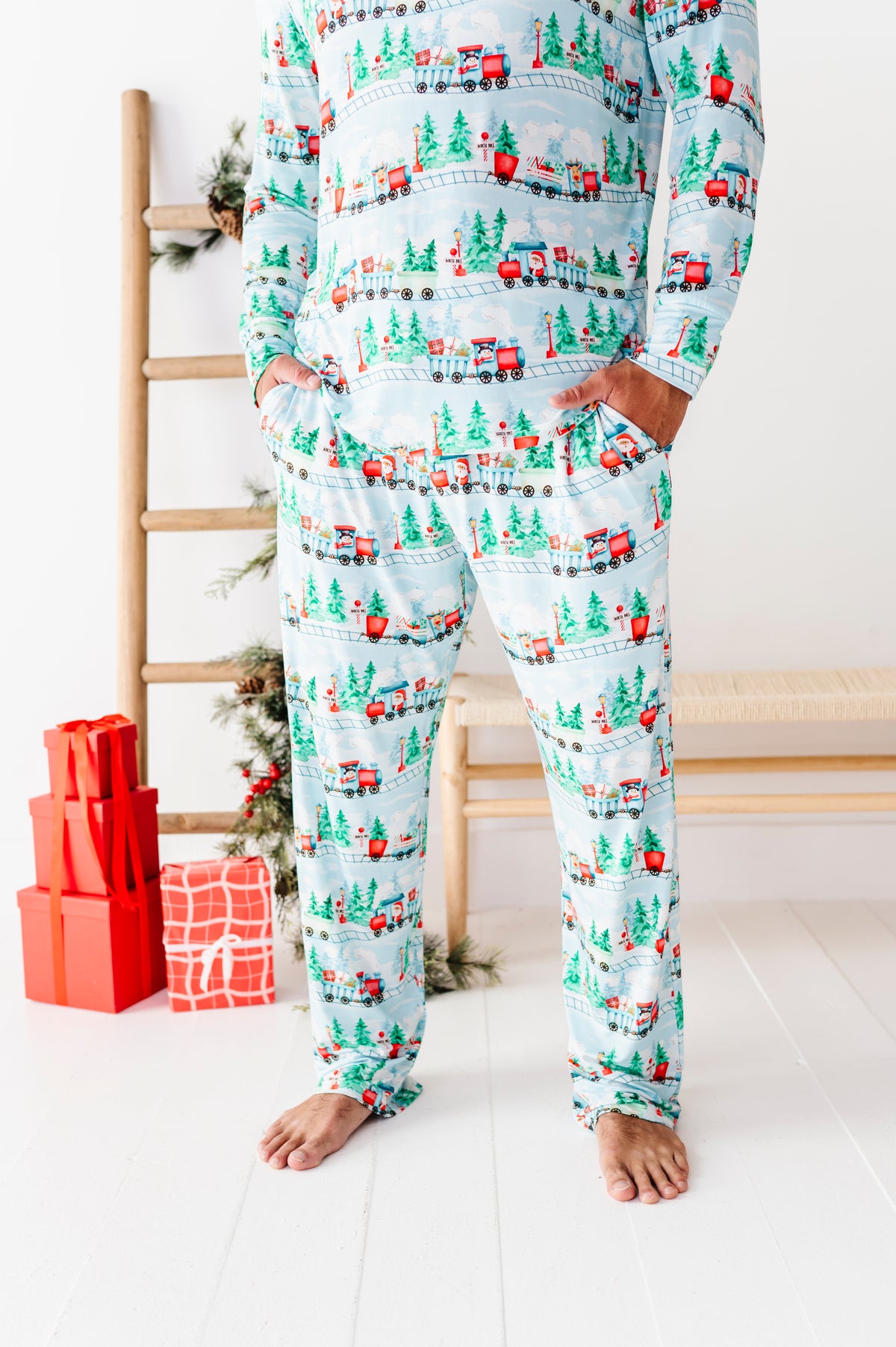 A man wearing a light blue long-sleeve bamboo pajama set with a festive Christmas print of red trucks, green trees, and holiday houses, standing barefoot in a bright room with wrapped gifts and a wooden ladder.