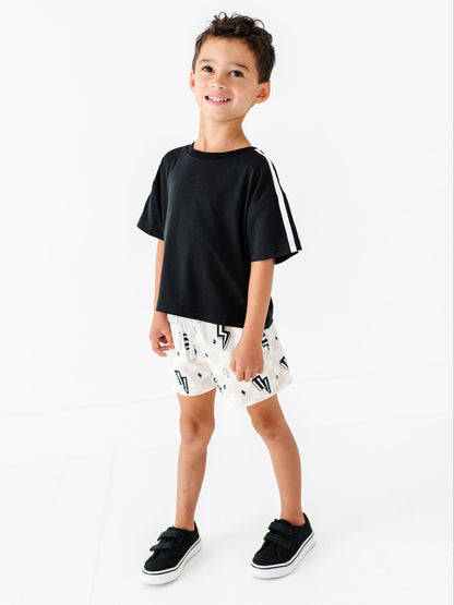 A young boy modeling a black short-sleeve t-shirt with white stripes on the sleeve, paired with white shorts featuring a black graphic pattern, and black slip-on shoes, set against a plain white studio background.