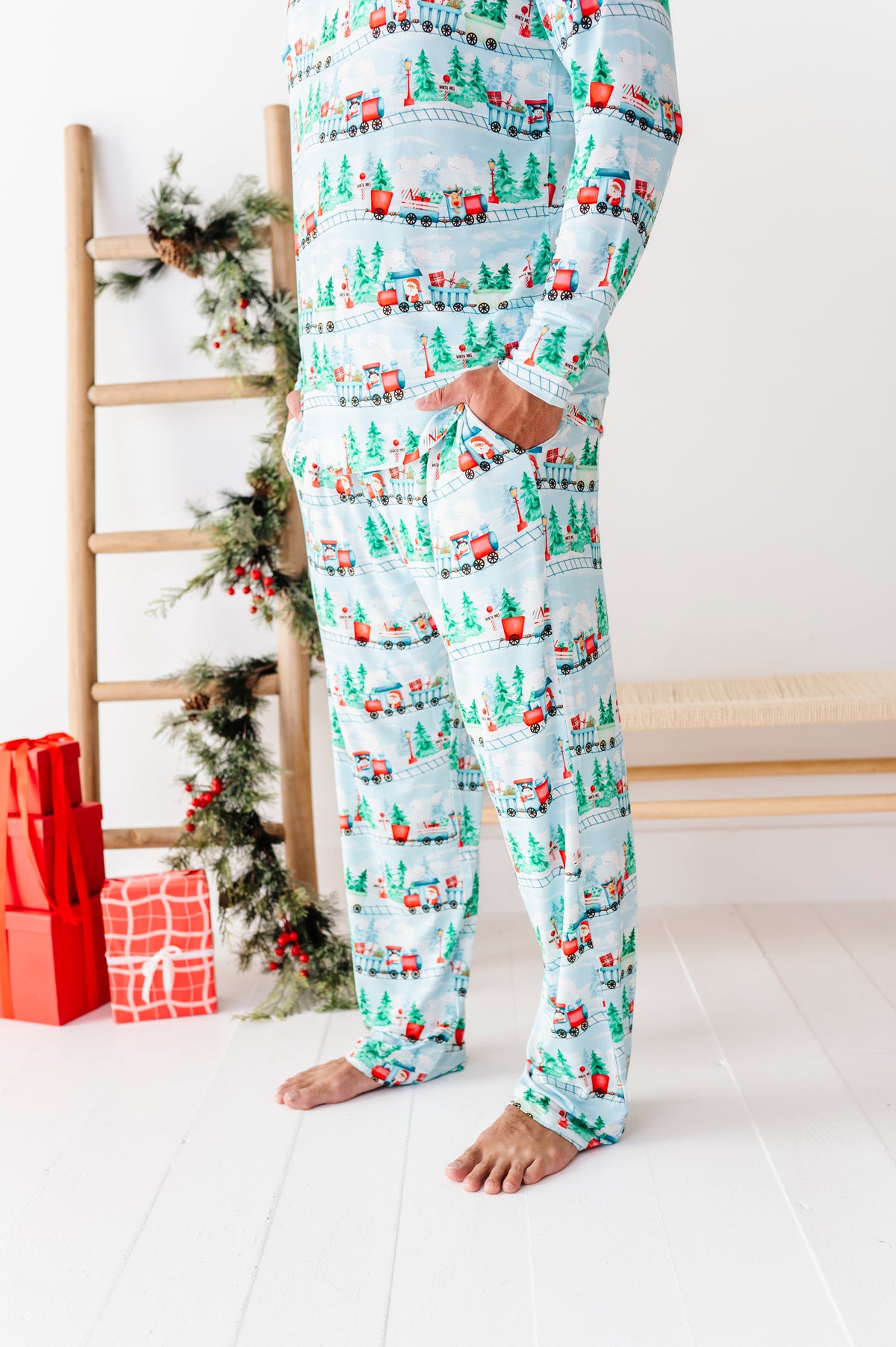 A dad wearing a light blue long-sleeve bamboo pajama set with a Christmas Santa Claus train print, standing barefoot in a bright room with a wooden ladder and holiday decorations.