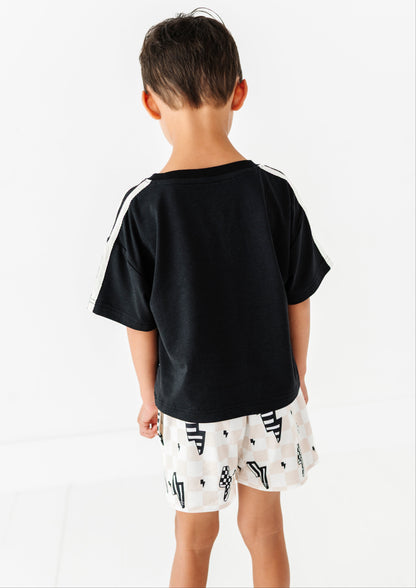 A child viewed from behind wearing a black short-sleeve t-shirt with white stripes on the shoulders and white shorts with a black geometric pattern, set against a plain white background.