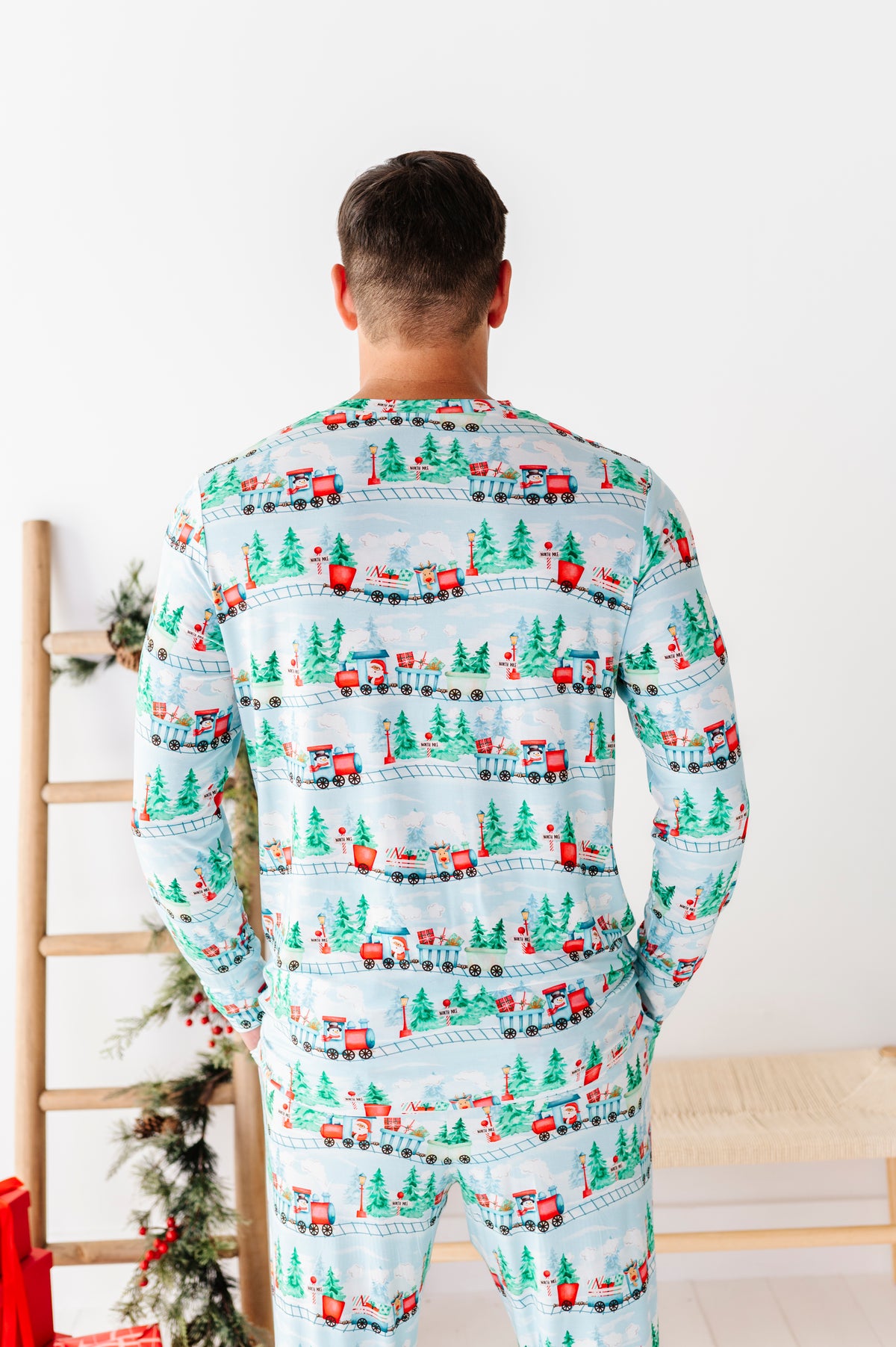 A man viewed from behind wearing a light blue long-sleeve bamboo pajama set with a repeating Christmas train pattern featuring red trains, green trees, and holiday motifs, set against a bright white room background.