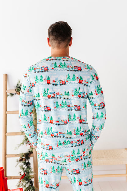 A man viewed from behind wearing a light blue long-sleeve bamboo pajama set with a repeating Christmas train pattern featuring red trains, green trees, and holiday motifs, set against a bright white room background.