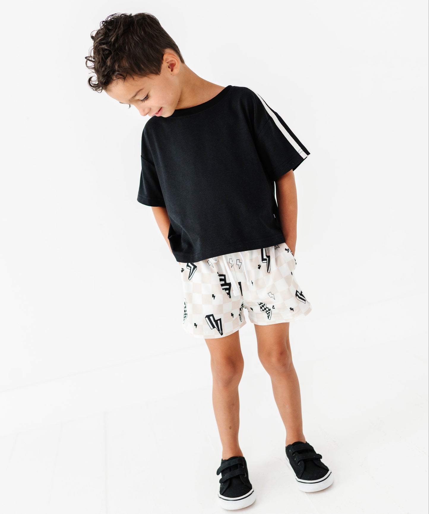 A young boy wearing a black short-sleeve t-shirt with white stripes on the sleeve, white shorts with a black graphic print, and black sneakers, standing against a plain white background.