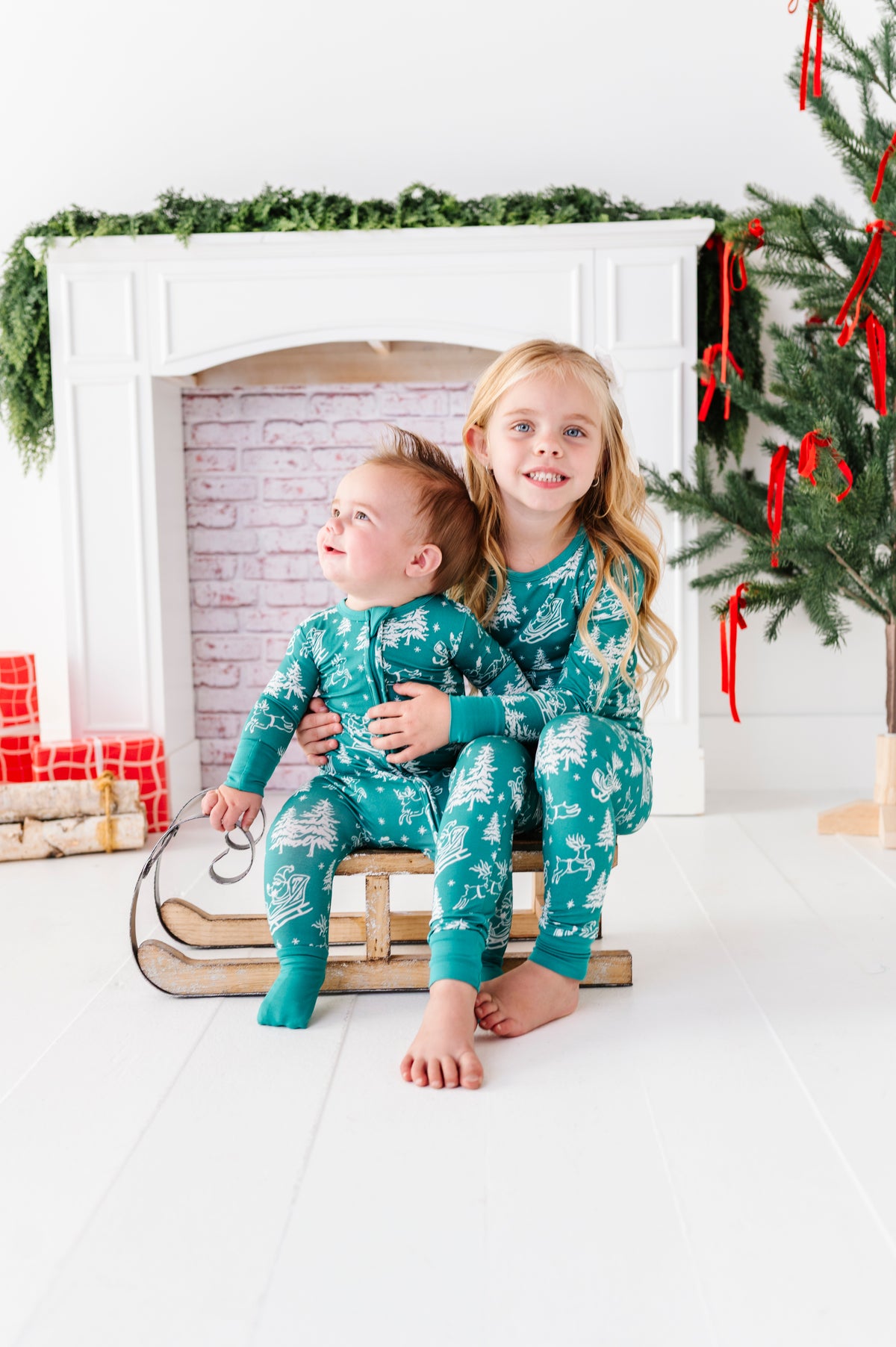 A brother and sister posed on a vintage sleigh. The brother and sister are wearing matching bamboo pajamas featuring vintage Santa and his reindeer 