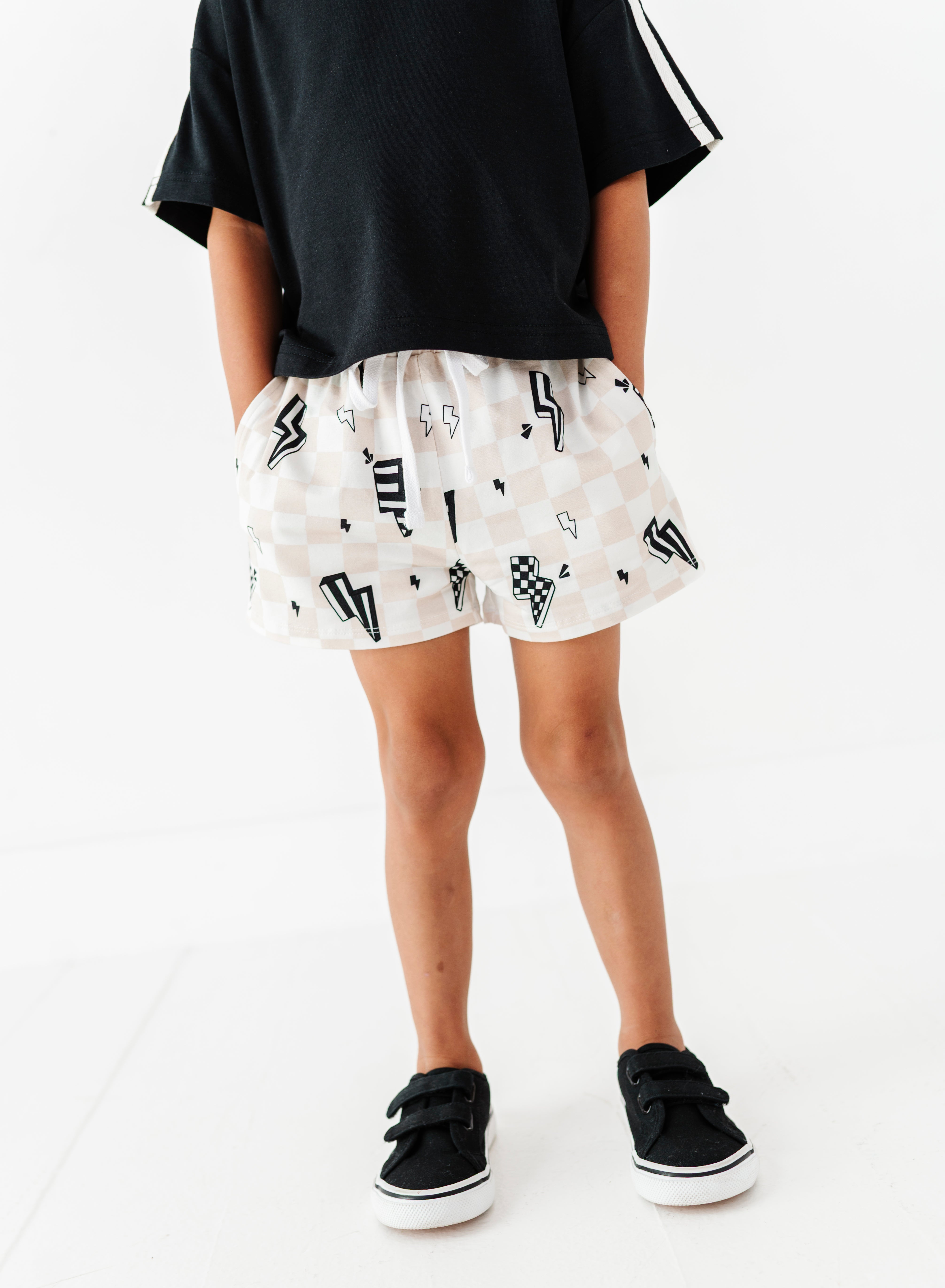 A child wearing white shorts with a black geometric pattern, a black t-shirt, and black sneakers, shown from the waist down against a white studio background.