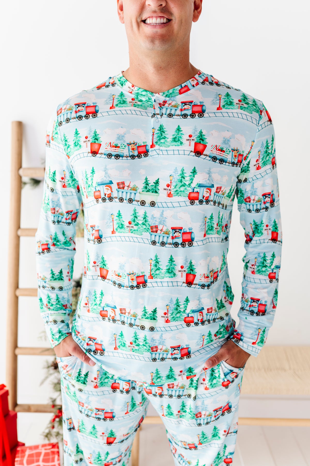 A man wearing a light blue long-sleeve bamboo pajama set with a repeating Christmas train and pine tree print, standing indoors with hands in pockets.