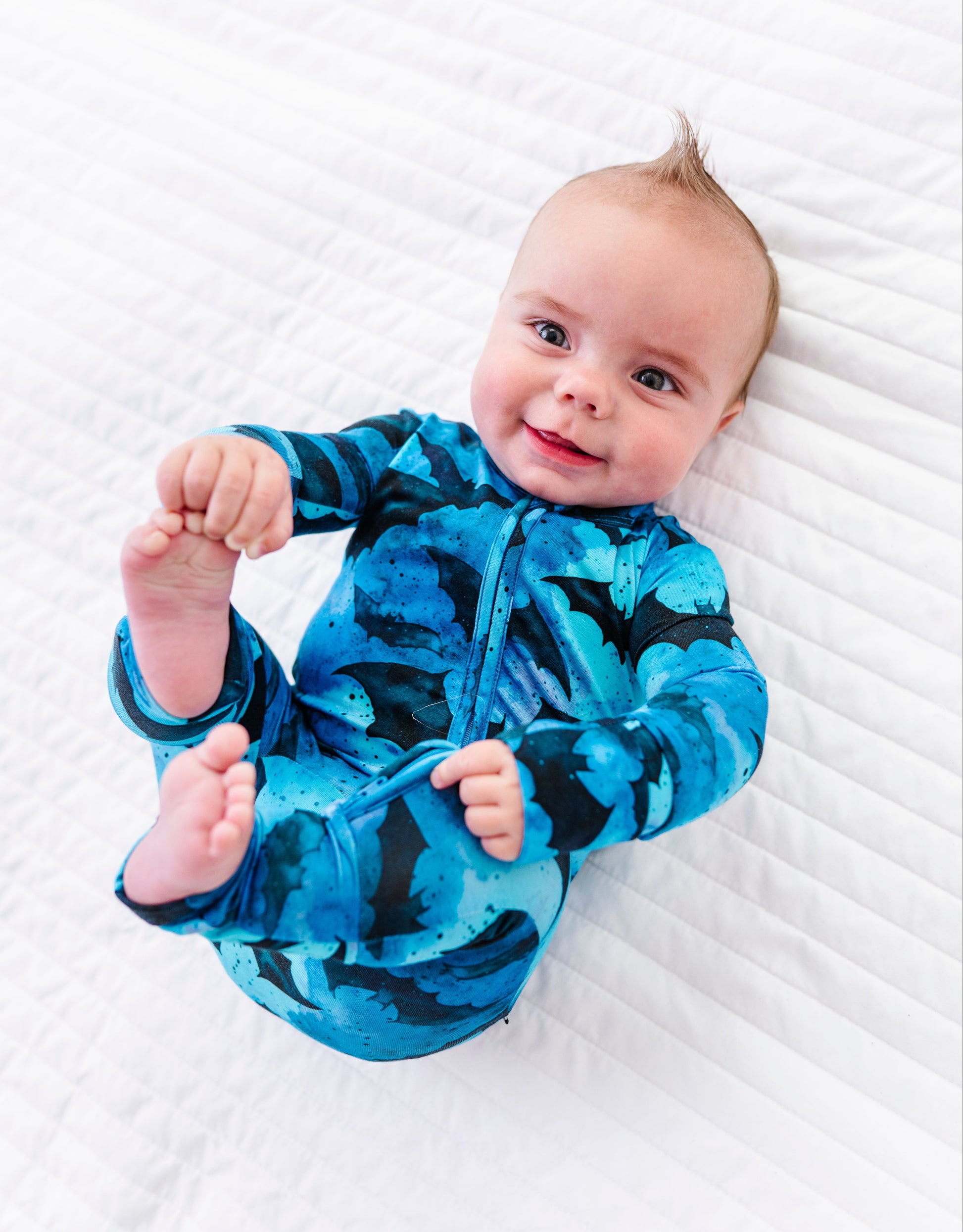 A baby lying on a white quilted blanket, wearing a blue and black patterned zip-up sleeper.