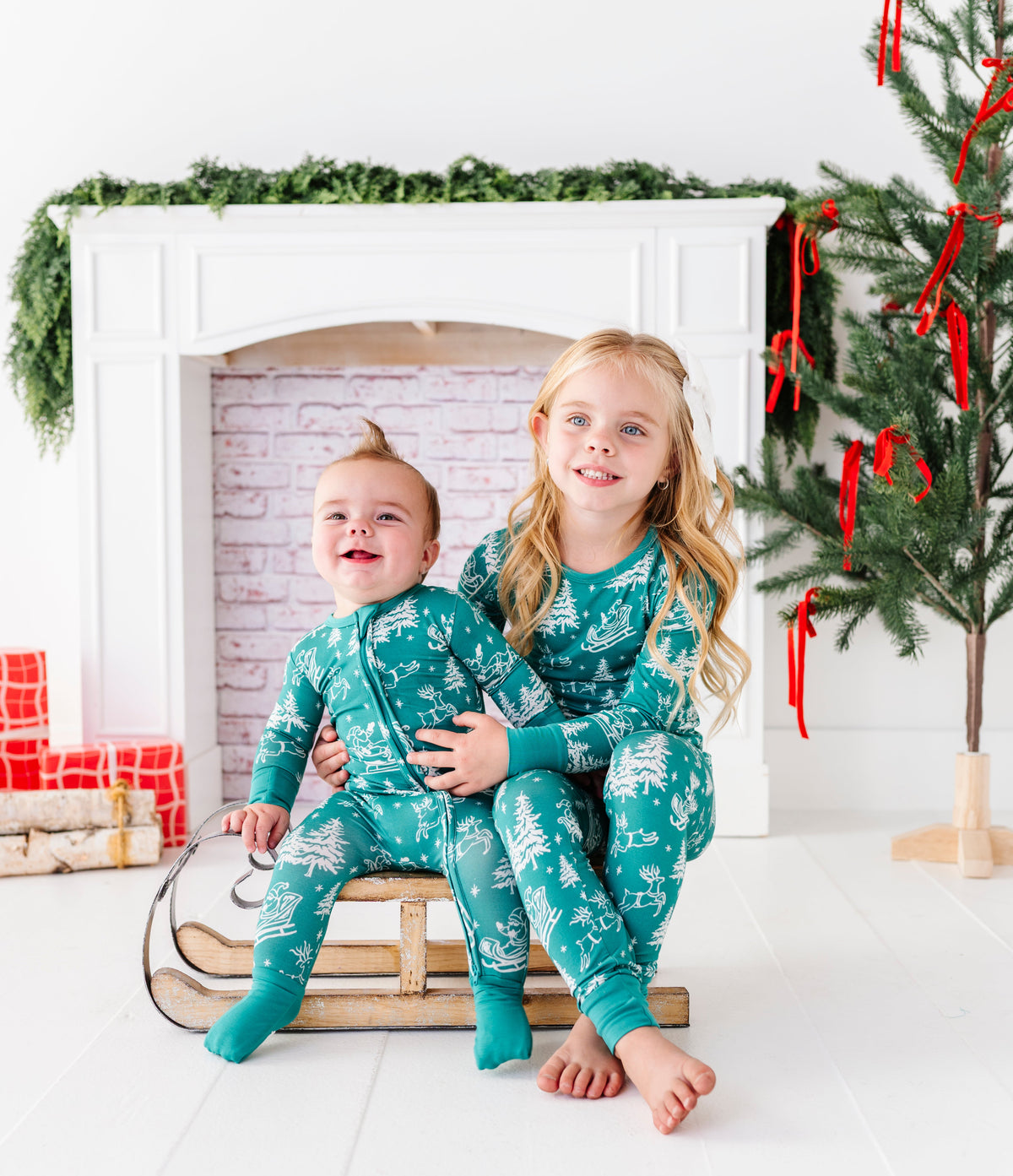 A brother and sister sitting on a wooden sleigh. They're wearing matching bamboo pajamas featuring vintage Santa and his reindeer 