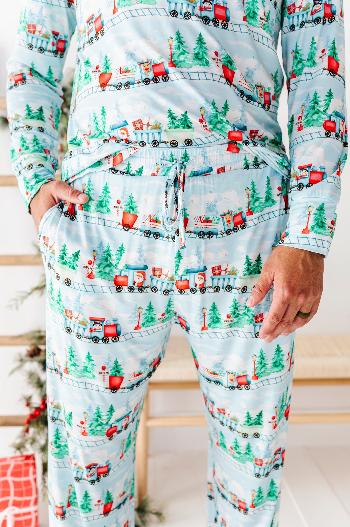 A person wearing a matching long-sleeve bamboo pajama set with a Christmas train and pine tree print on a light blue background, standing indoors with hands in the pockets.
