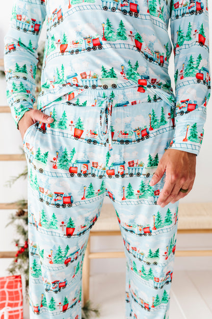 A person wearing a matching long-sleeve bamboo pajama set with a Christmas train and pine tree print on a light blue background, standing indoors with hands in the pockets.