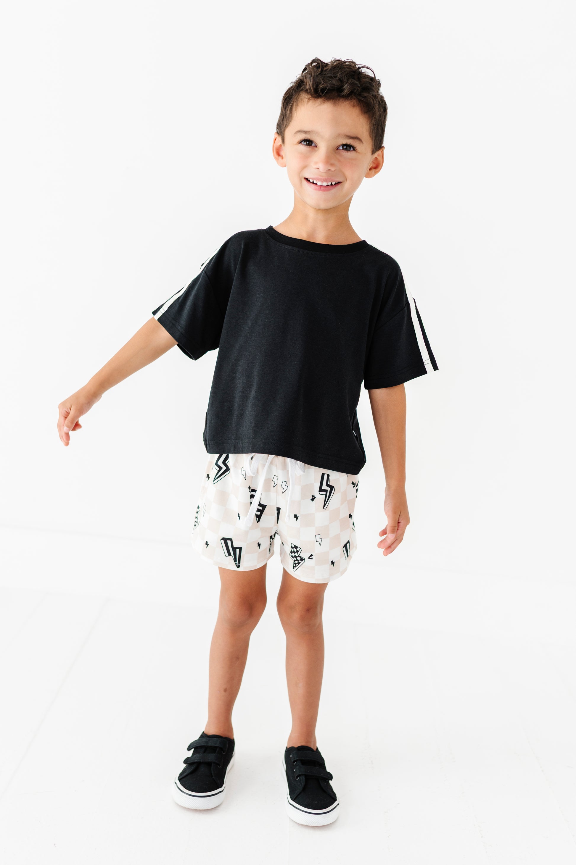 A young boy modeling a black short-sleeve t-shirt with white stripes on the sleeves, paired with white shorts featuring a black lightning-bolt pattern, and black sneakers, set against a plain white studio background.