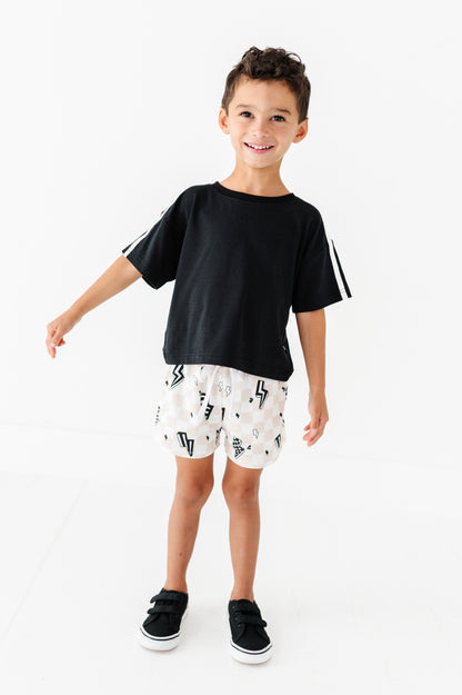 A young boy modeling a black short-sleeve t-shirt with white stripes on the sleeves, paired with white shorts featuring a black lightning-bolt pattern, and black sneakers, set against a plain white studio background.