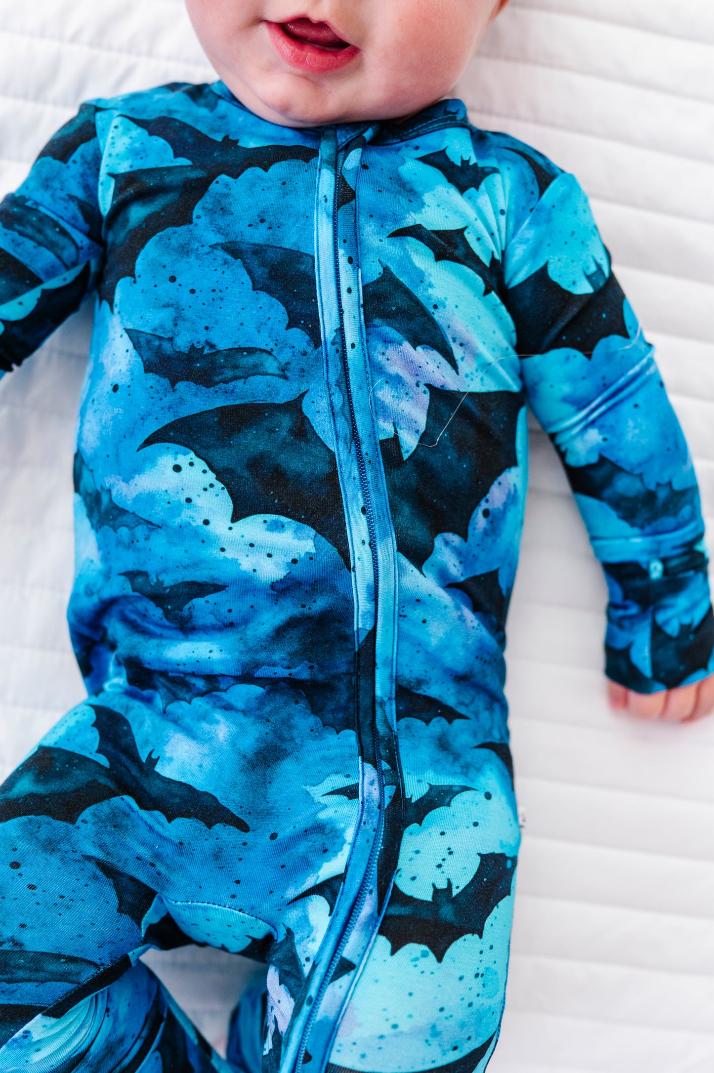 A close-up of a baby wearing a blue and black bat-patterned zip-up sleeper, lying on a white quilted mattress.