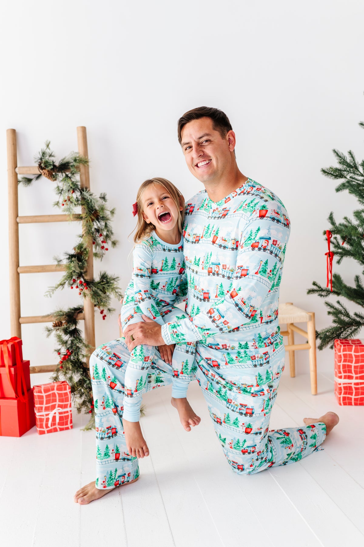 A man and a young girl wearing matching Christmas-themed bamboo pajamas with a snowman and tree pattern, posing together in a decorated holiday room.