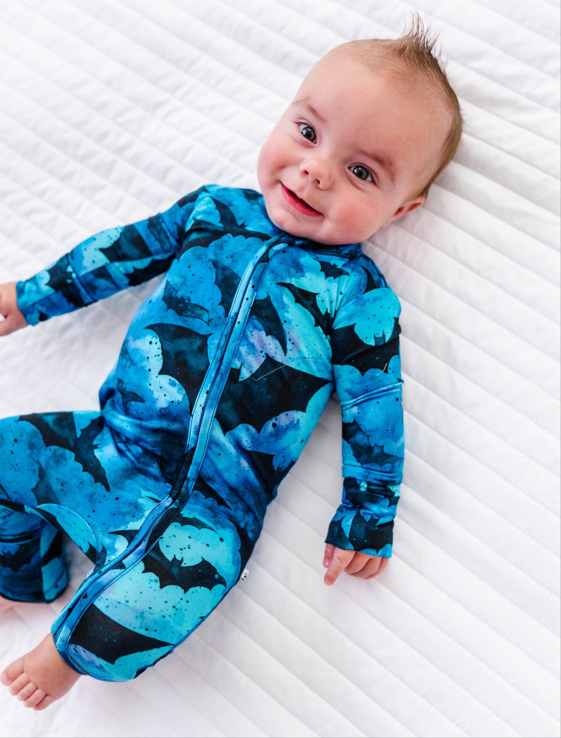 A baby lying on a white quilted blanket, wearing a long-sleeve footless onesie with a blue and black butterfly pattern.