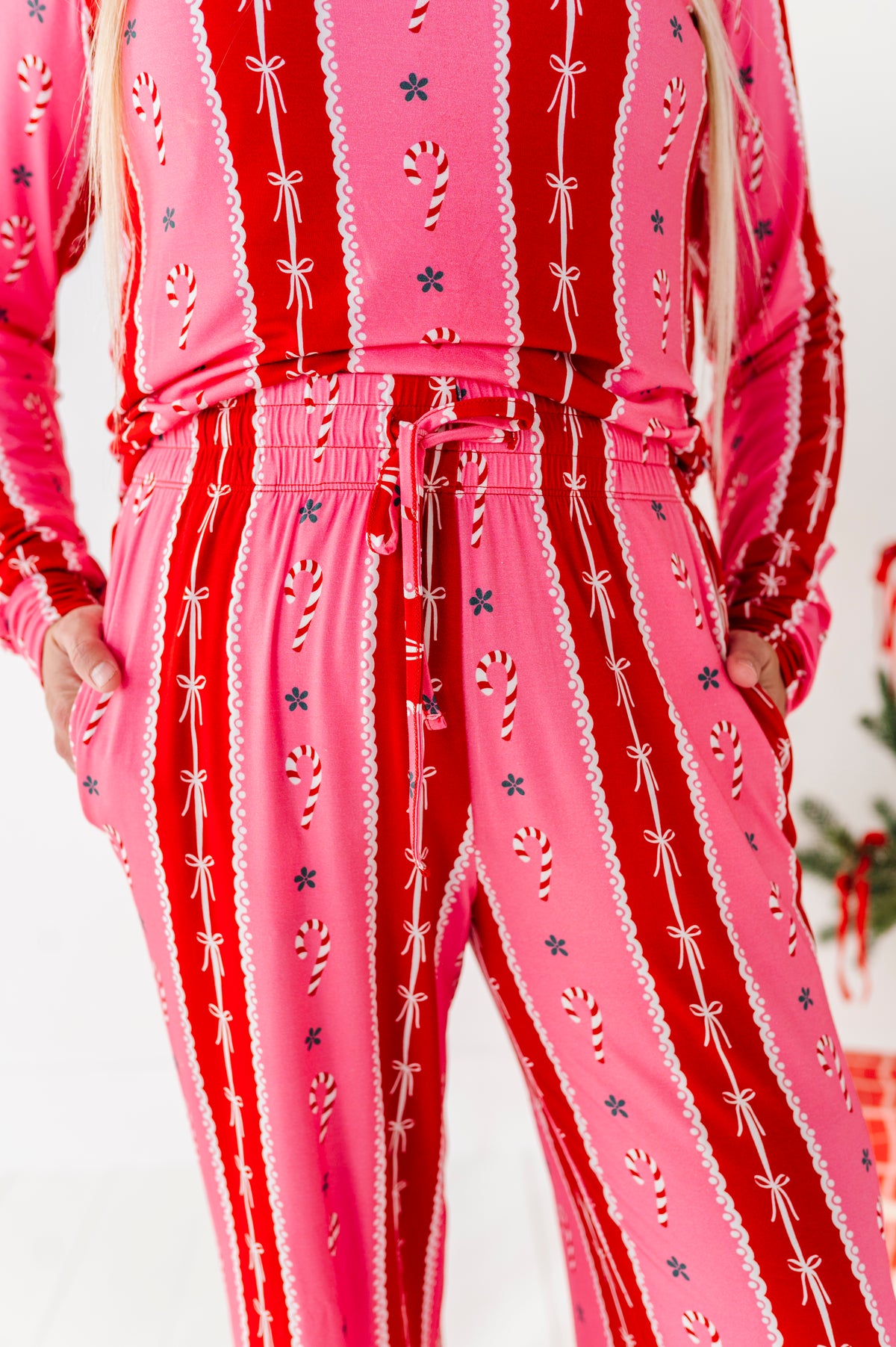 A person wearing a matching pink and red holiday-themed bamboo lounge set with vertical stripes, candy cane motifs, and white lace-like trim details. The photo is cropped from the chest down, showing the outfit against a light studio background with a small festive decoration at the far right edge.