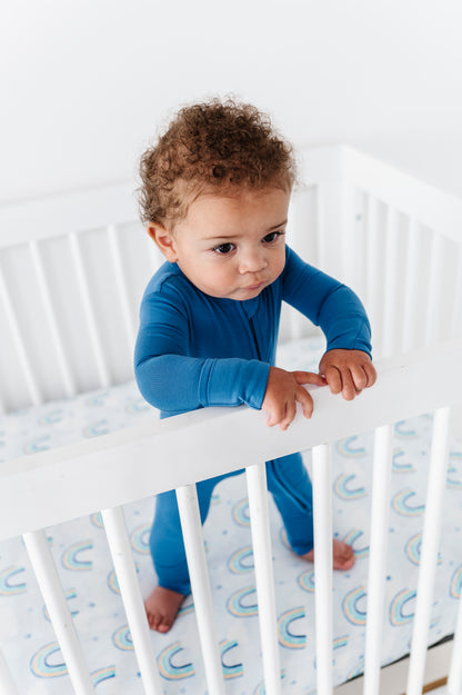 Child in a blue bamboo zip romper standing in a crib with a white and blue patterned bamboo crib sheet.