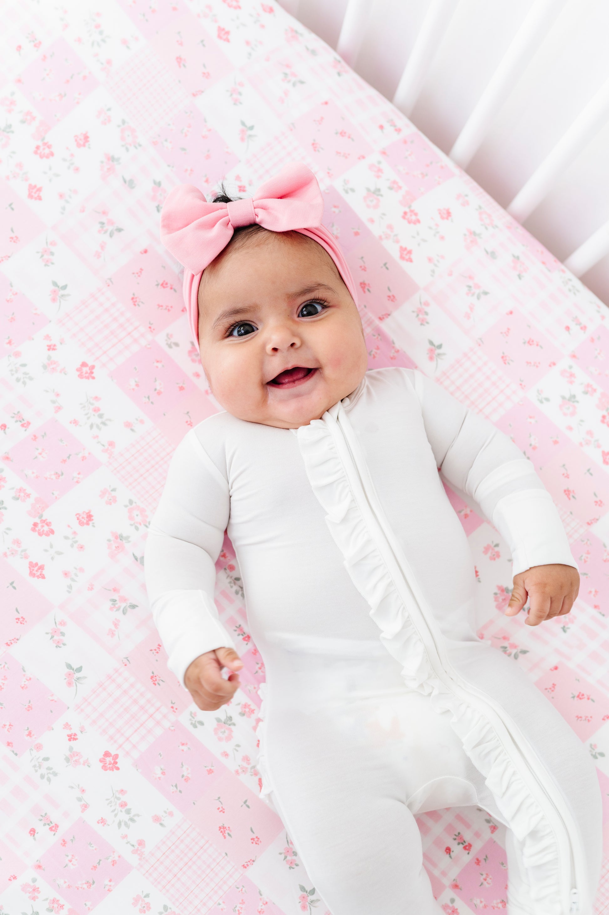 Baby in a white onesie with a pink bow headband on a floral bamboo crib sheet.