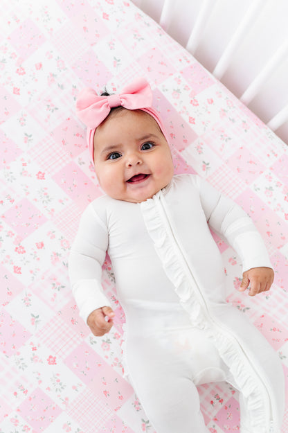 Baby in a white onesie with a pink bow headband on a floral bamboo crib sheet.