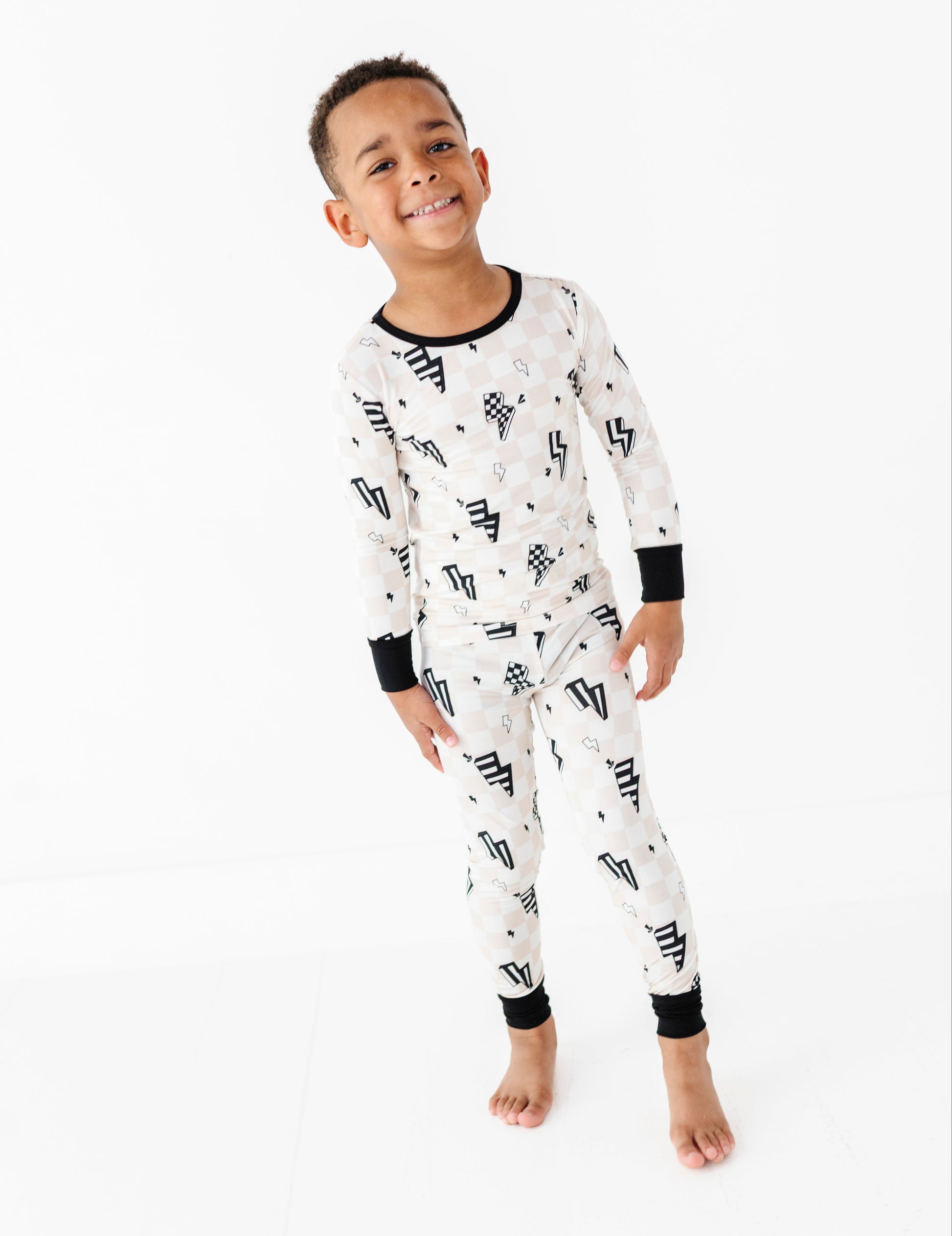 A young boy smiling and wearing a long-sleeve white pajama set with a black geometric pattern, photographed against a seamless white studio background.