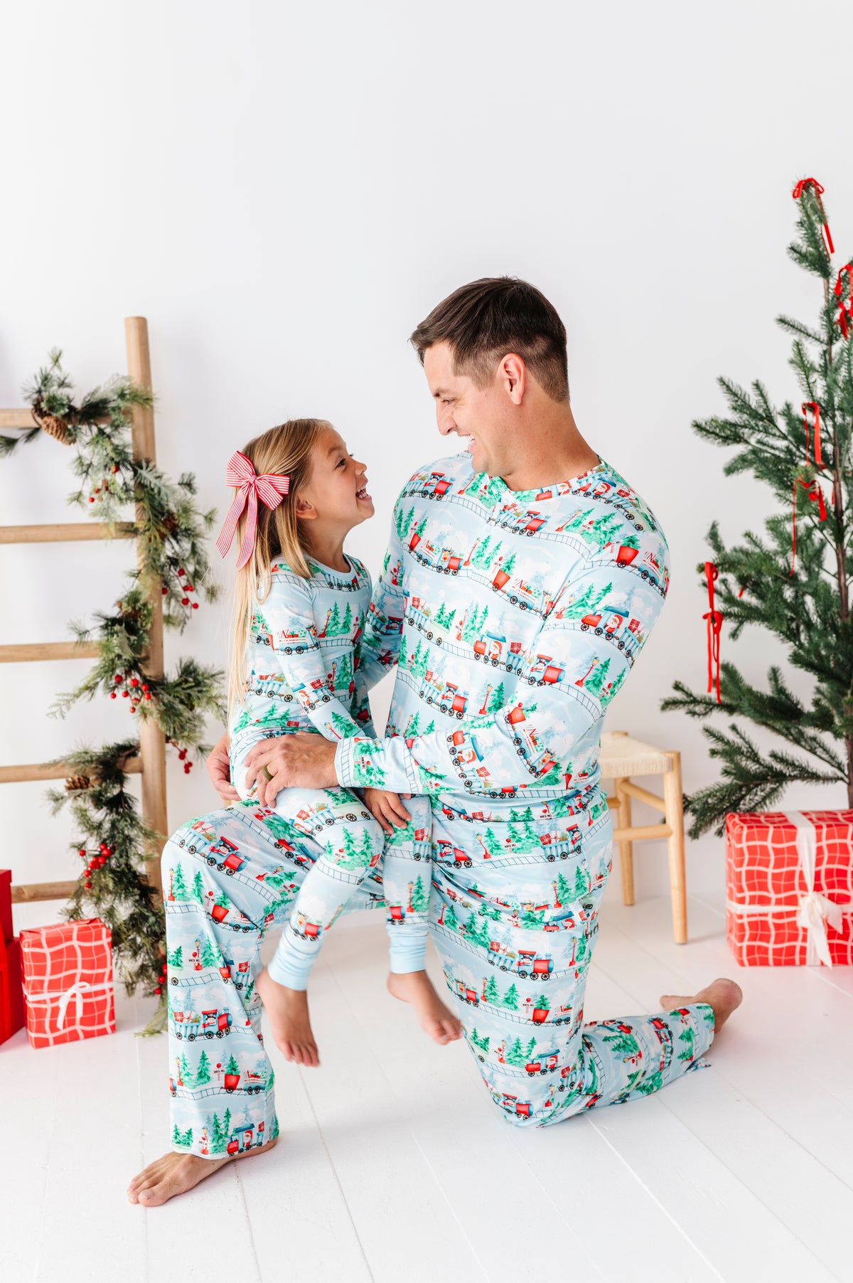A father and daughter wearing matching Christmas-themed bamboo pajamas with a green pine tree and red ornaments pattern, posed together in a brightly lit holiday room with decorated Christmas trees and wrapped gifts.