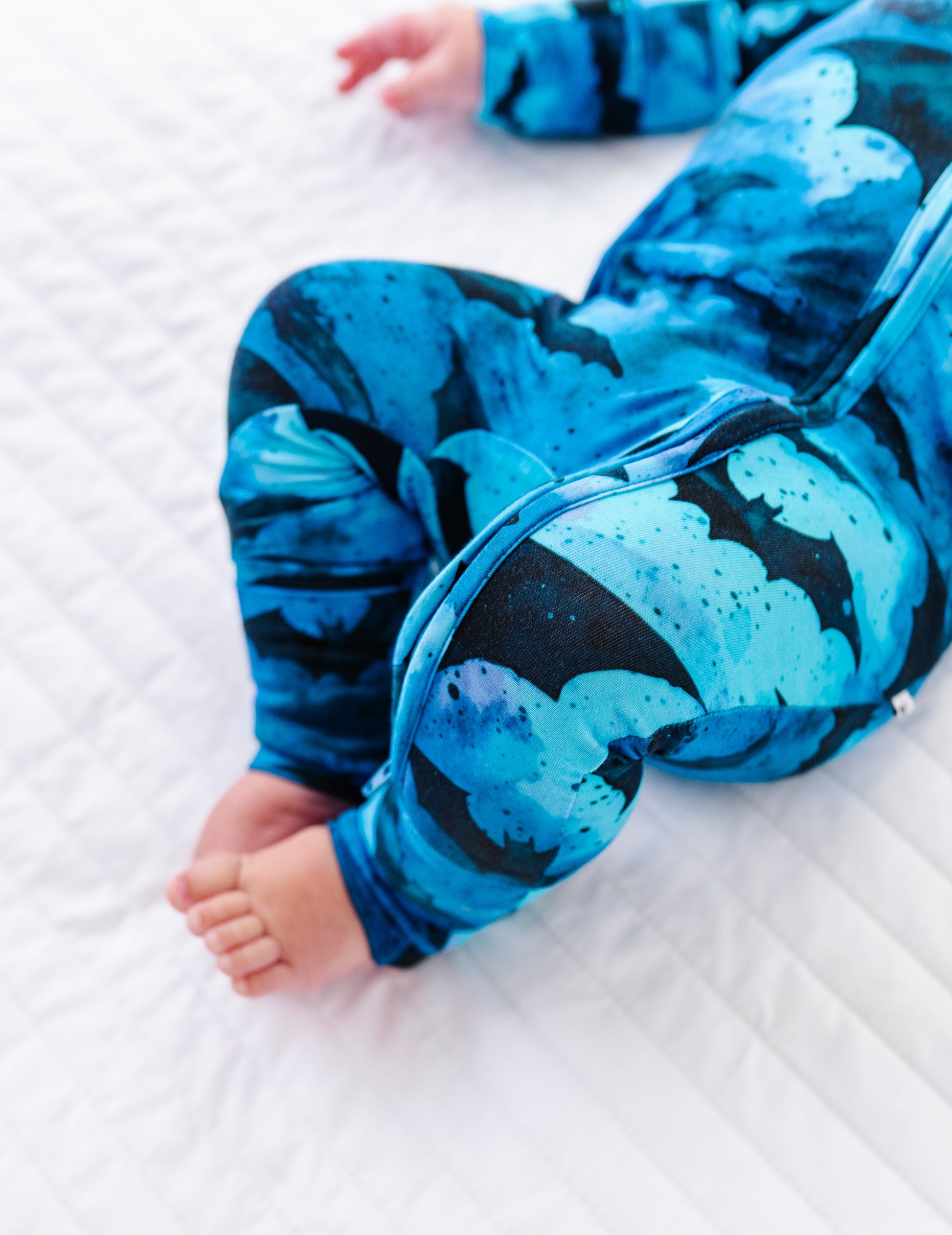 A baby wearing a blue and black abstract patterned one-piece jumpsuit lying on a white quilted mattress.
