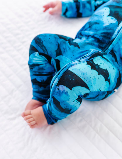 A baby wearing a blue and black abstract patterned one-piece jumpsuit lying on a white quilted mattress.