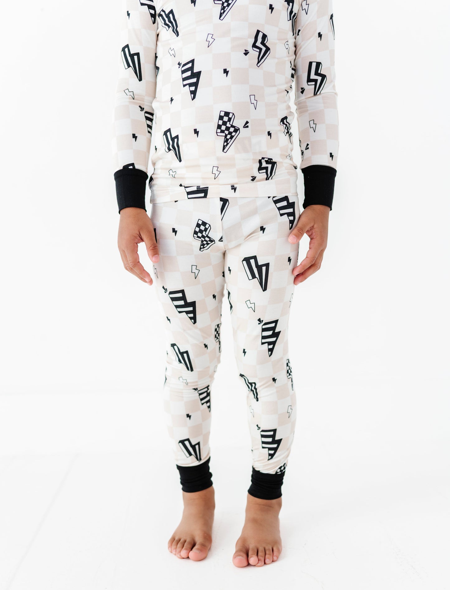 A child wearing a matching long-sleeve pajama top and pants set with a black and white checkerboard pattern, black lightning bolt motifs, and black ribbed cuffs, shown against a plain white studio background.