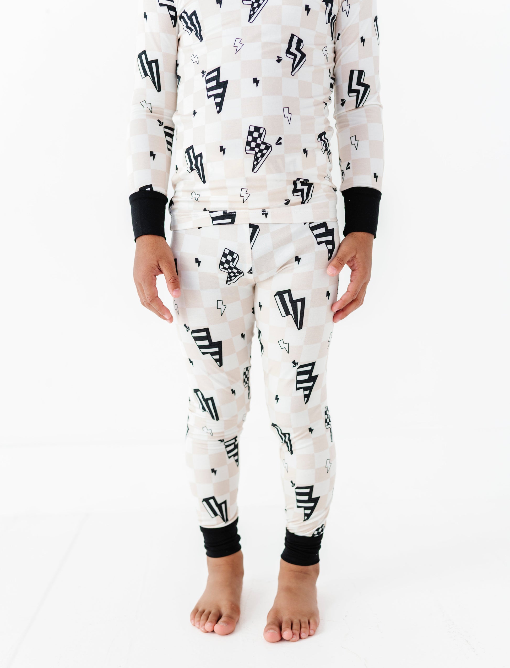 A child wearing a matching long-sleeve pajama top and pants set with a black and white checkerboard pattern, black lightning bolt motifs, and black ribbed cuffs, shown against a plain white studio background.