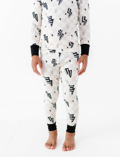 A child wearing a matching long-sleeve pajama top and pants set with a black and white checkerboard pattern, black lightning bolt motifs, and black ribbed cuffs, shown against a plain white studio background.