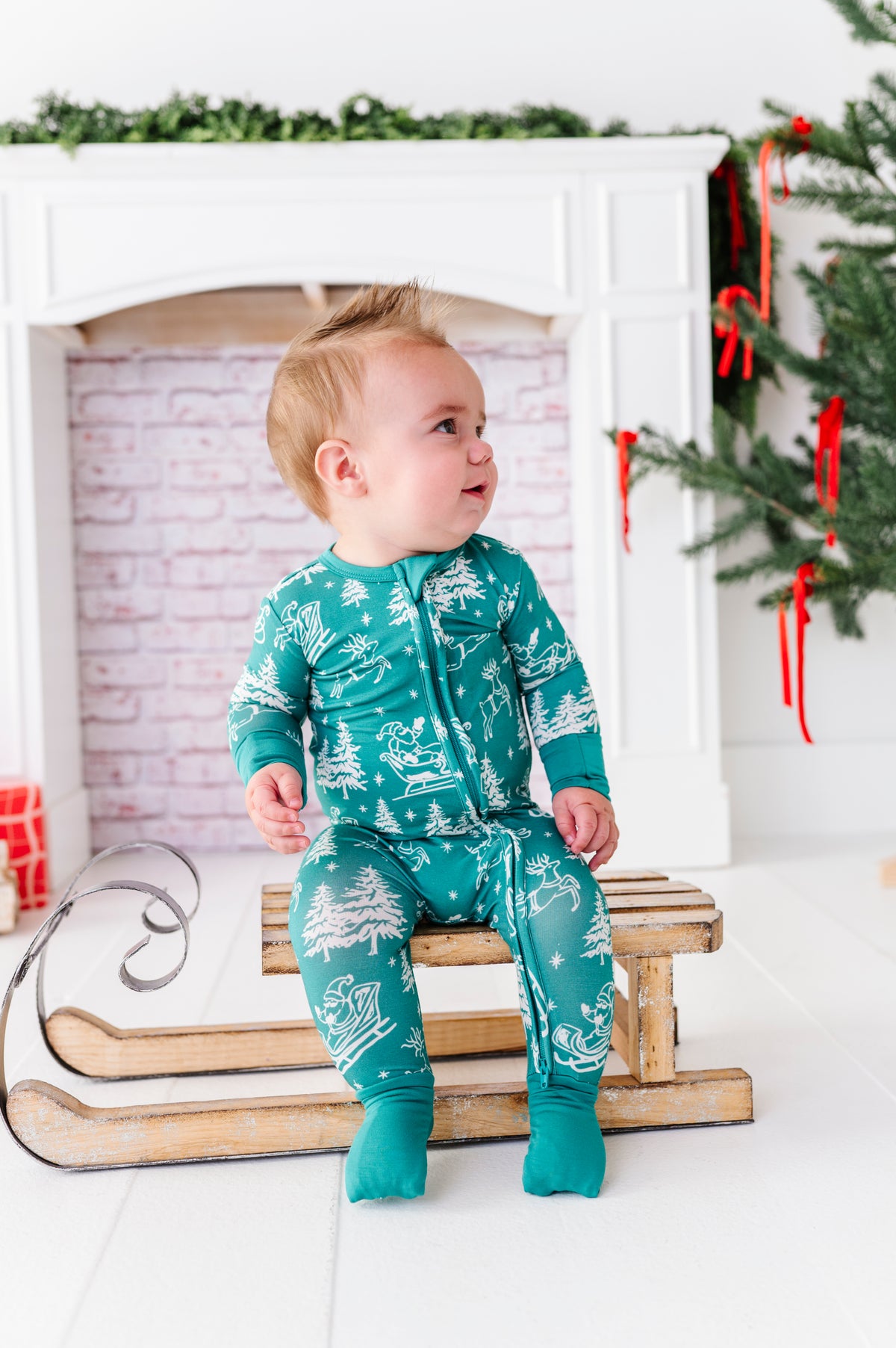 A little boy sitting on an old wooden sleigh. He's wearing a bamboo convertible romper featuring vintage scenery including Santa and his reindeer 