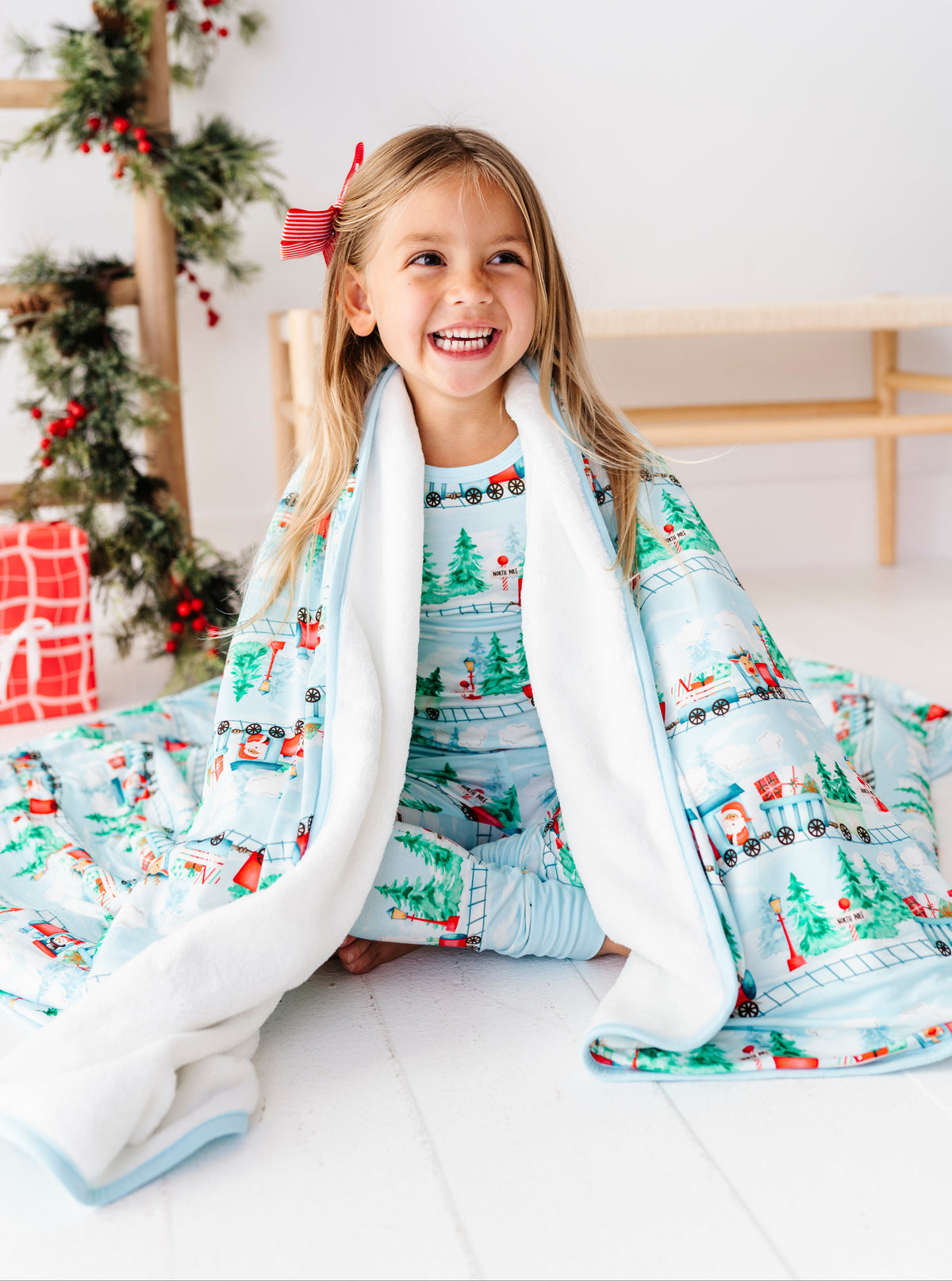 A young girl smiling while sitting on a white floor, wrapped in a light blue Christmas-themed blanket and matching pajamas. The blanket features a festive print of snowmen, Christmas trees, and trains, with a soft white sherpa lining.