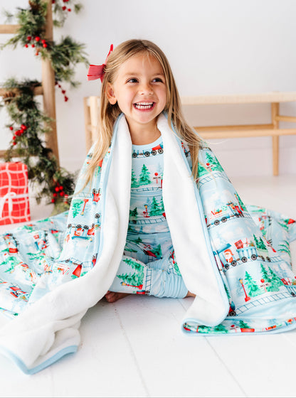 A young girl smiling while sitting on a white floor, wrapped in a light blue Christmas-themed blanket and matching pajamas. The blanket features a festive print of snowmen, Christmas trees, and trains, with a soft white sherpa lining.