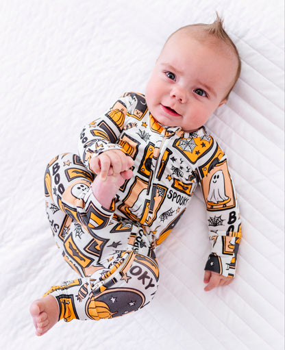 A baby lying on a white quilted blanket, wearing a long-sleeve footed sleeper with a Halloween-themed graphic print in yellow, black, and white.