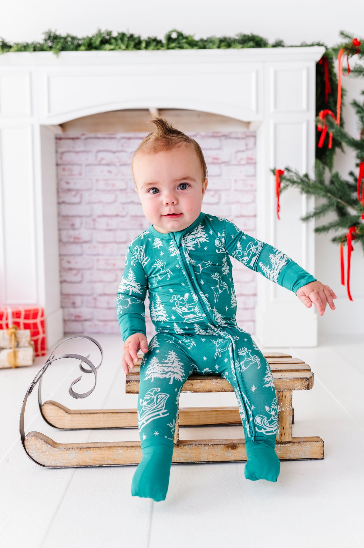 A little boy sitting on a wooden sleigh, wearing a bamboo convertible romper featuring vintage scenery of Santa and his reindeer 