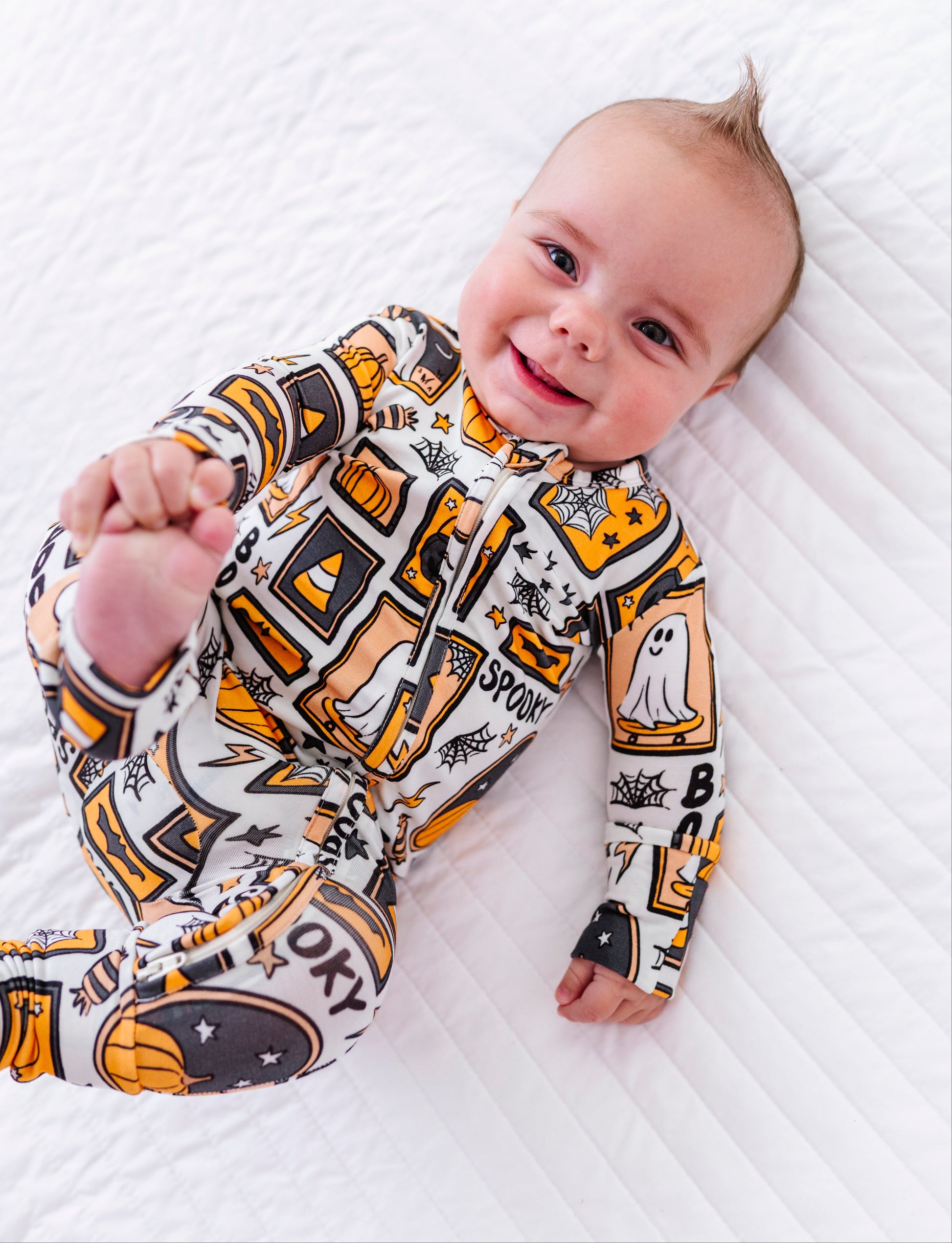 A baby lying on a white quilted blanket, wearing a long-sleeve footed sleeper with a Halloween-themed graphic print in orange, black, and white.